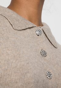 Beige wollen trui met een kraaghalslijn, voorzien van drie decoratieve metalen knopen met rhinestones. Geverfd breisel.