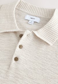 Beige strukturiertes Poloshirt mit einem gerippten Kragen, ausgestattet mit drei braunen Knöpfen und einem Etikett. Das Material wirkt weich und leicht.