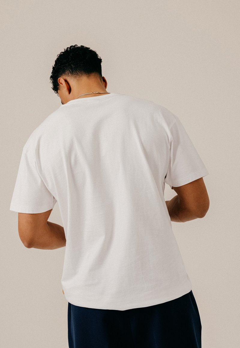 Homme aux cheveux courts et bouclés, portant un t-shirt blanc uni à manches courtes et un pantalon sombre, de dos sur un fond neutre.