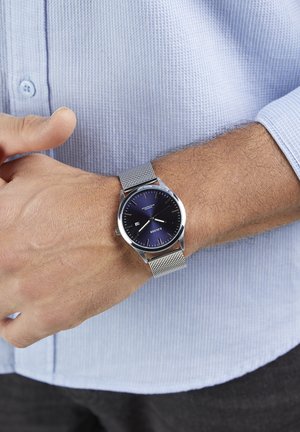 Round watch with a navy blue dial, silver hour markers, and a silver mesh strap, worn on a wrist with short brown hair.
