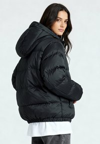 Veste matelassée noire avec capuche, présentant un design capitonné, un ourlet élastiqué, et une texture lisse et brillante. Le modèle est montré de dos.