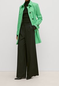 Un trench-coat léger vert avec des boutons noirs et des poignets ceints, associé à un pantalon noir taille haute à jambes larges et des escarpins noirs à bout pointu.