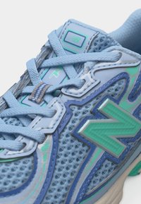 Κοντινό πλάνο ενός αθλητικού παπουτσιού με μπλε διχτυωτό υλικό, λογότυπο New Balance σε τόνους τιρκουάζ και μοβ, γαλάζια κορδόνια και λεπτομερές ράψιμο στην πλευρά και στη γλώσσα του παπουτσιού.