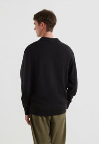 Maglione nero a maglia con colletto, con vestibilità comoda e polsini a costine, abbinato a pantaloni khaki chiari. La texture sembra morbida.