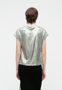 Άτομο με κοντά καστανά μαλλ�ιά, φορώντας ένα λαμπερό ασημένιο μπλουζάκι κοντού μανικιού και μαύρη φούστα, στέκεται με την πλάτη στραμμένη προς την κάμερα.