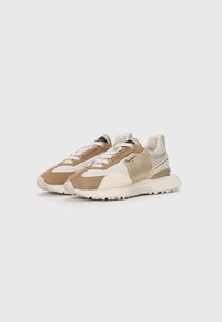 Sneakers featuring a mix of tan, beige, and cream suede and fabric, with a white sole and contrasting black accent on the tongue.