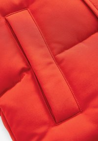 Gilet matelassé rouge vif en tissu lisse et texturé, avec une poche rectangulaire et des coutures visibles. Conception compacte et isolante.