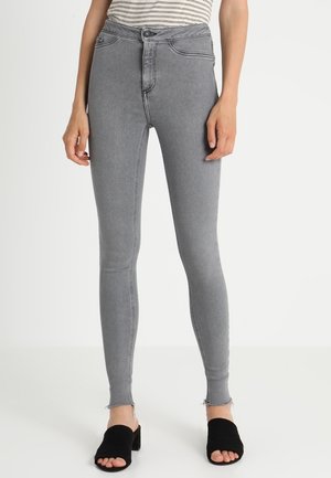 Kobieta w szarych rurkach z wysokim stanem z postrzępionymi brzegami oraz czarnych, otwartych na palce sandałach na słupku, stojąca na białym tle.