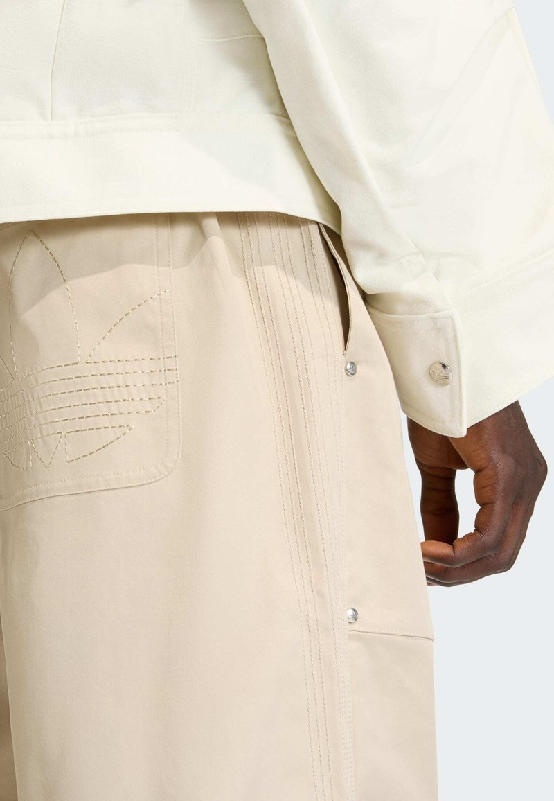 Cream jacket with a close-fitting collar and beige pants featuring an embroidered logo. The pants have decorative stitching and snap fasteners.