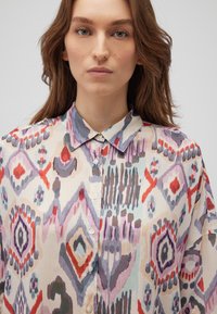 Patterned blouse in lightweight, semi-sheer fabric featuring geometric designs in purple, red, and blue hues; collared with button closures.
