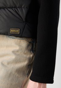Colete puffer preto com camisa de manga longa preta canelada, jeans de ganga e um rótulo bege com "Barbour International" em dourado.