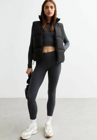 Chaleco negro acolchado, camiseta de manga larga gris, mallas grises acanaladas y zapatillas blancas con detalles en marrón. Texturas suaves, diseño ajustado.