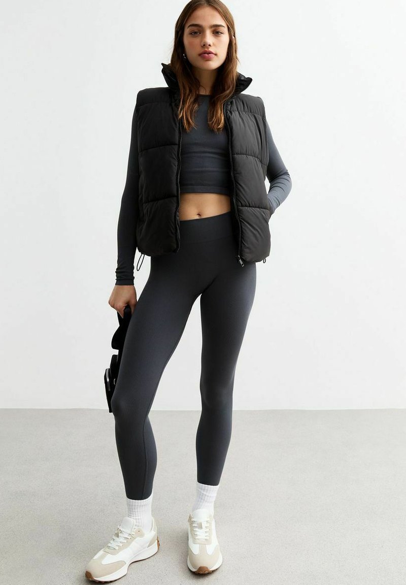 Chaleco negro acolchado, camiseta de manga larga gris, mallas grises acanaladas y zapatillas blancas con detalles en marrón. Texturas suaves, diseño ajustado.