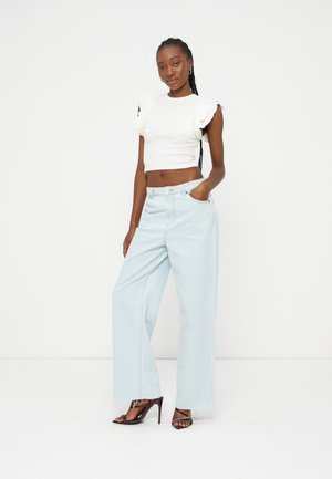 Femme aux cheveux tressés portant un haut court blanc sans manches à volants, un jean large bleu clair et des sandales noires à talons hauts, debout.