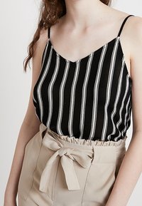 Black and white striped camisole with thin straps, featuring a loose fit and soft fabric, paired with high-waisted beige trousers with a bow.