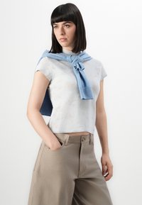 Lyseblå blomstermønstret strikket topp med korte ermer, sammen med beige vide bukser; en blå genser bundet løst rundt skuldrene.