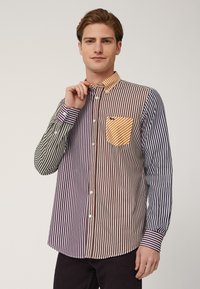 Camicia a righe con colori alternati di viola, marrone, verde e blu/navy. Ha una chiusura a bottoni sul davanti, un colletto a contrasto e una tasca sul petto.