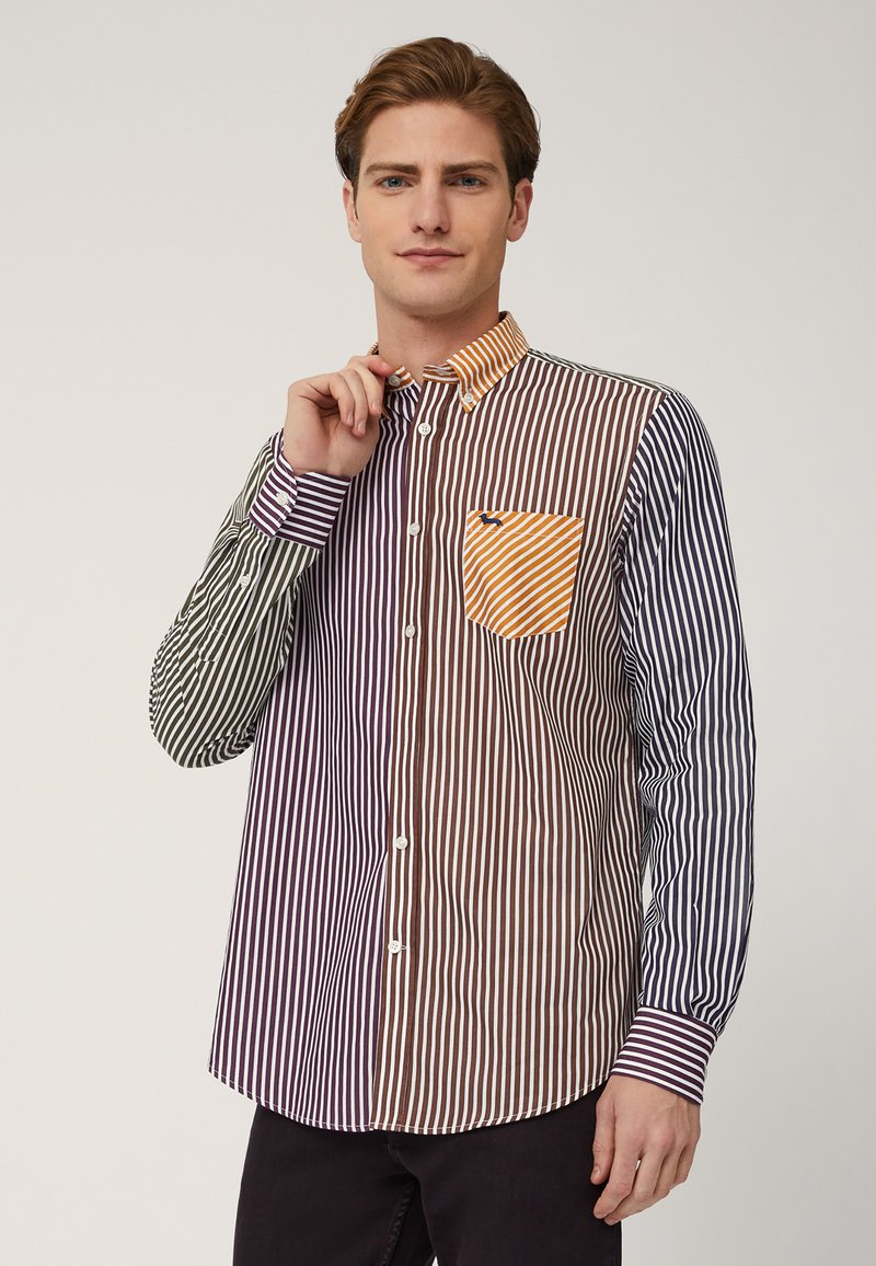 Camicia a righe con colori alternati di viola, marrone, verde e blu/navy. Ha una chiusura a bottoni sul davanti, un colletto a contrasto e una tasca sul petto.