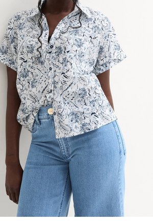 Donna che indossa una camicia bianca a maniche corte con motivo floreale blu e jeans azzurri a vita alta.