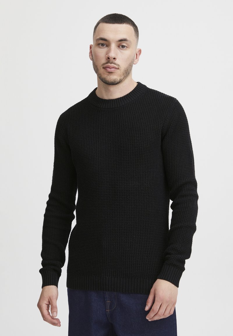 Black knitted sweater with a ribbed crew neckline, textured fabric, fitted sleeves, and a straight hem, paired with denim pants.