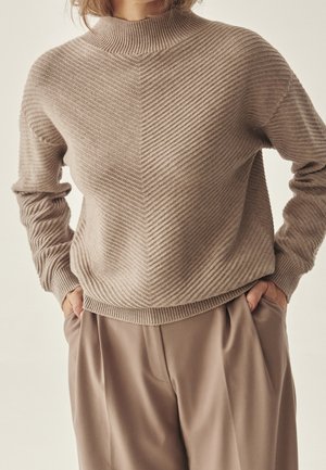 Person trägt einen gerippten, taupefarbenen Rollkragenpullover und weite taupefarbene Hose, steht mit den Händen in den Taschen vor einem schlichten Hintergrund.