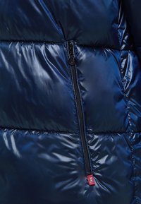 Granatowa puchowa kurtka o błyszczącej, pikowanej fakturze. Posiada czarny zamek błyskawiczny oraz małą czerwoną metkę z logo w pobliżu zamka.