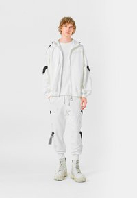Sudadera blanca con cremallera y acentos bordados en negro, camiseta blanca, pantalones de chándal a juego y botas de combate gris claro. Textura suave.