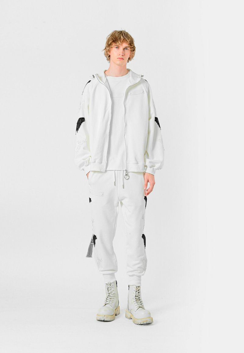 Sudadera blanca con cremallera y acentos bordados en negro, camiseta blanca, pantalones de chándal a juego y botas de combate gris claro. Textura suave.
