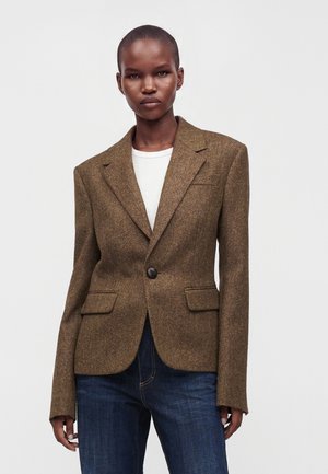 Femme aux cheveux courts porte un blazer marron ajusté avec un bouton, une chemise blanche et un jean bleu sur un fond blanc uni.