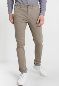 Man i beiga slim-fit byxor, grå långärmad skjorta och vita sneakers står mot en vit bakgrund.