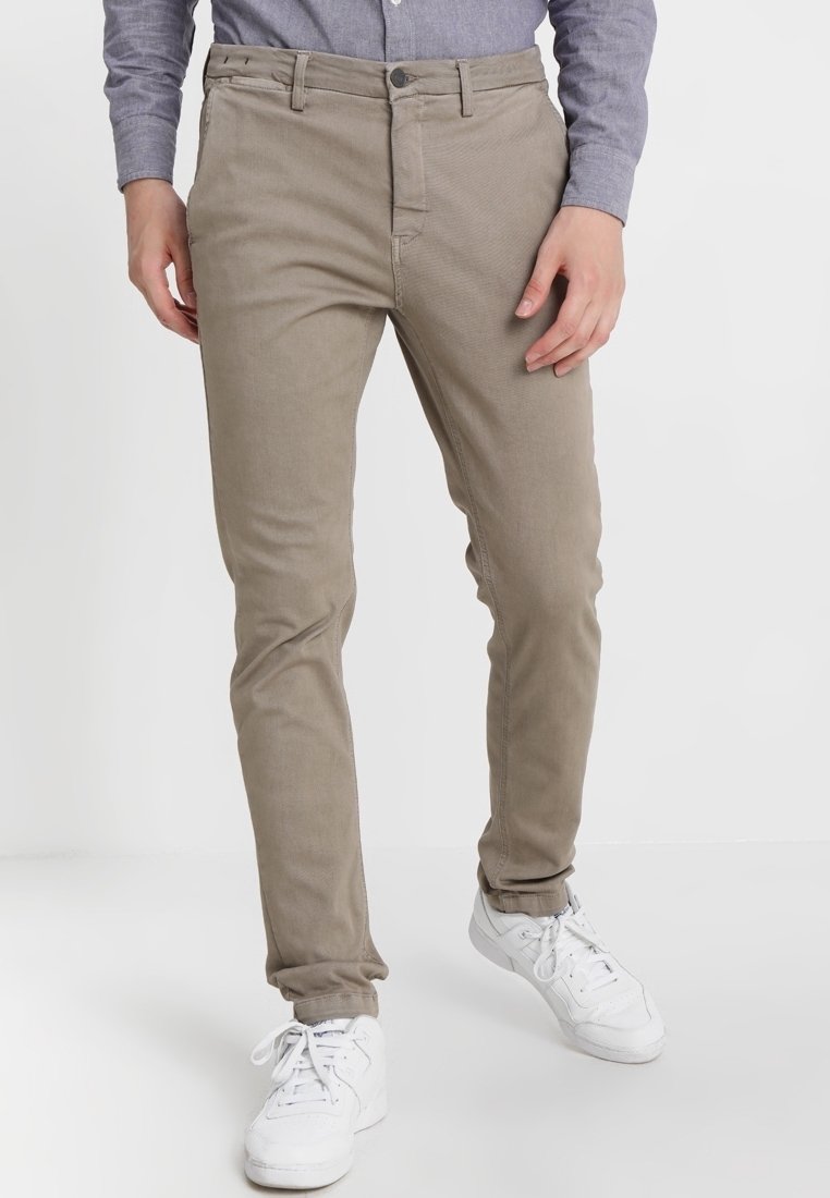 Man i beiga slim-fit byxor, grå långärmad skjorta och vita sneakers står mot en vit bakgrund.