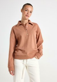 Svetle hnědý polokošilový svetr ze směsi bavlny s límečkem, středním zapínáním na knoflíky a pletenými rukávy, doplněný o bílé kalhoty.