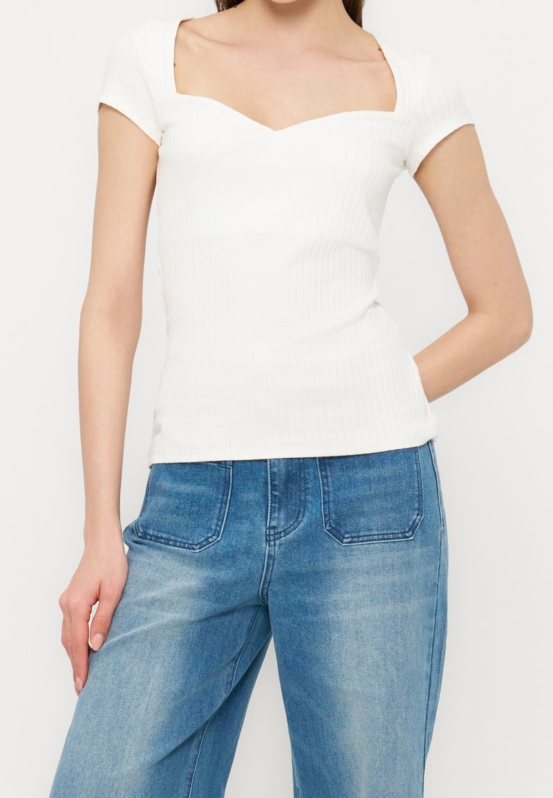 Femme portant un top blanc ajusté à manches courtes avec un décolleté en cœur et un jean en denim bleu taille haute avec poches avant.