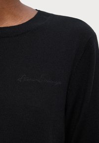 Maglione nero lavorato a maglia con scollo a girocollo e logo ricamato discreto sul petto. Texture liscia con una vestibilità aderente.