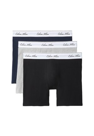 Jonge man zonder shirt die zwarte boxershorts met witte tailleband draagt, staat op blote voeten tegen een simpele grijze achtergrond.