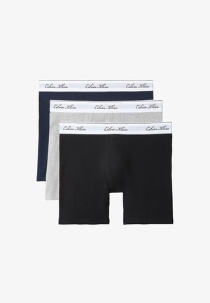 Jonge man zonder shirt die zwarte boxershorts met witte tailleband draagt, staat op blote voeten tegen een simpele grijze achtergrond.