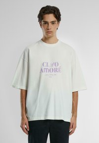 Overdimensjonert hvit bomull t-skjorte med korte ermer, med en lilafarget grafisk trykk som leser "Ciao Amore" i midten.