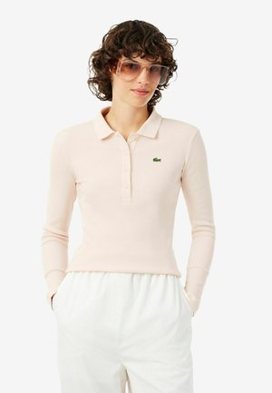 Jeune personne aux cheveux bouclés portant de grandes lunettes de soleil translucides, un polo à manches longues beige clair avec logo, et un pantalon blanc les mains dans les poches.