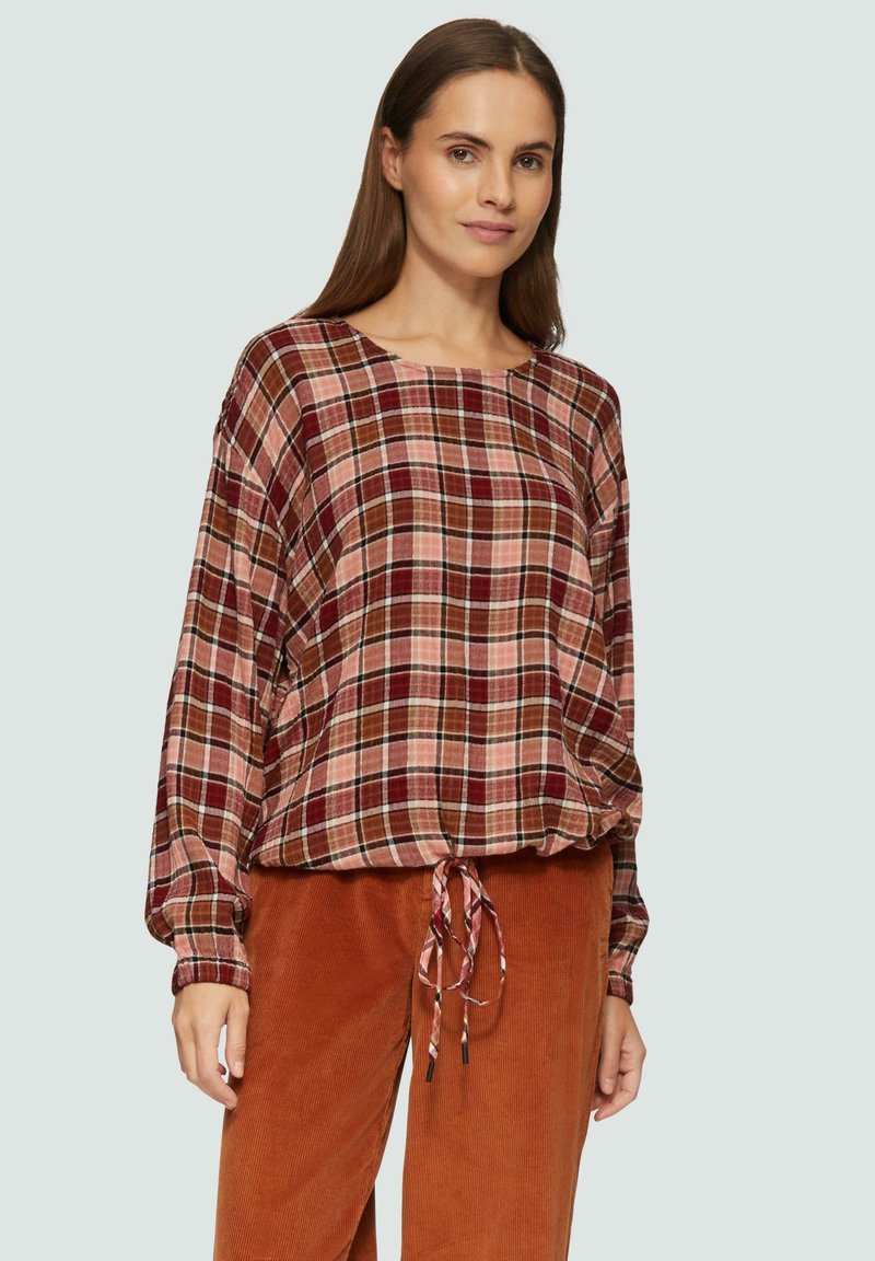 Chemisier à manches longues à carreaux dans des teintes de rouge, marron et rose, avec un col rond et un lien ajustable à l'ourlet. Associé à un pantalon en velours côtelé.
