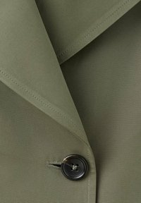 Gros plan sur un tissu vert olive avec un bouton noir cousu près de la couture du revers d’un vêtement sur mesure.