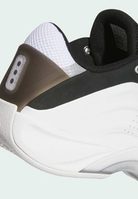 Scarpa sportiva bianca e nera con linguetta in rete testurizzata, suola in gomma e un accento marrone lucido sul tallone con dettagli perforati.