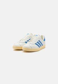 Zapatillas blancas con acentos azules. Presentan tres rayas azules en los laterales, suela de goma texturizada y un collar acolchado para mayor soporte.
