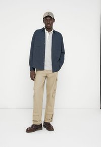 Μπλε σκούρο πουκάμισο με κουμπιά, ανοιχτό γκρι πόλο, μπεζ παντελόνια cargo, καφέ παπούτσια με κορδόνια και μπεζ καπέλο, εμφανίζονται πάνω σε λευκό φόντο.