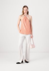 Ferskenfarget paljetthaltertop, hvite vide bukser med sidespalter, og en liten rosa håndveske. Modell stående mot en enkel bakgrunn.