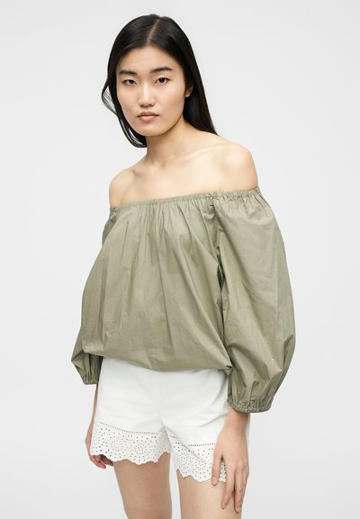 Γυναίκα με μακριά μαύρα μαλλιά φοράει χαλαρό ελιάς πράσινο τοπ χωρίς ώμους και λευκά σορτς με δαντέλα, ποζάροντας μπροστά σε απλό ανοιχτό υπόβαθρο.