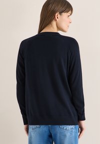 Femme aux cheveux bruns raides portant un pull à manches longues bleu marine et un jean bleu clair, vue de dos sur un fond beige uni.
