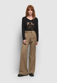 Roodharige vrouw staat in een zwarte top met lange mouwen en een metalen grafisch ontwerp, een beige gestreepte wijde broek met tailleband en zwarte schoenen.