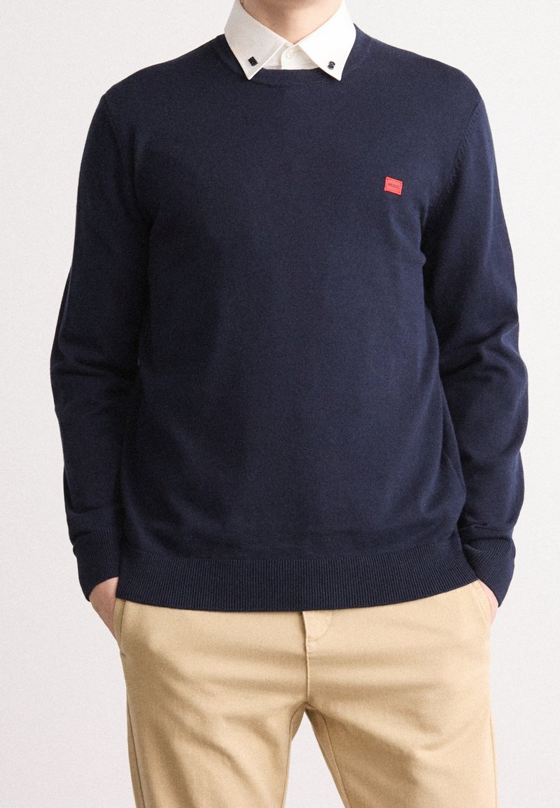 Marinblå tröja i mjukt stickat material, med rund halsringning och en liten röd logotyp på vänster bröst.