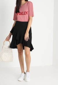 T-shirt rosa con la scritta "DARLIN'" in rosso, abbinata a una gonna nera con volant. Tiene in mano una borsa di paglia bianca. Indossa sneakers bianche.
