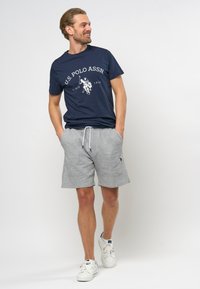 Tamno plavi pamučni majica sa bijelim logotipom "U.S. Polo Assn." i grafikom. Sivi pamučni šorc s elastičnom vrpcom i izvezenim logotipom. Bijele tenisice.